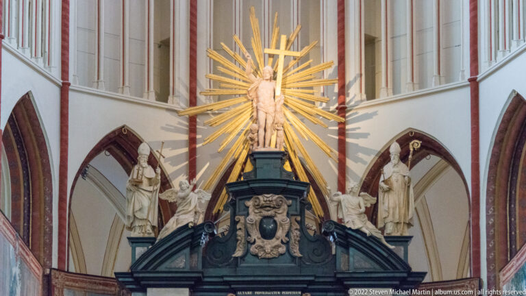 Crowned female sacred figure before a gilded sunburst above stone saints and heraldic ornament in the cathedral.