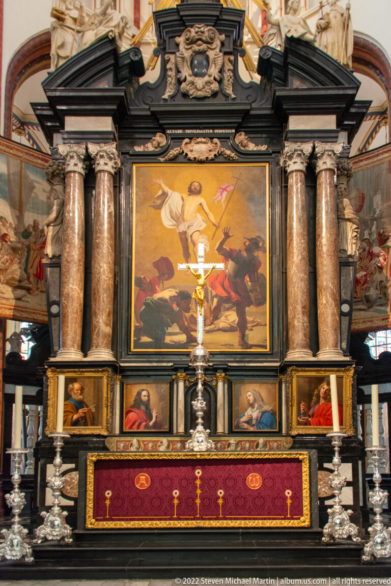 Cathedral altar with central religious painting, marble columns, silver candlesticks, crucifix, and red gold-embroidered frontal.