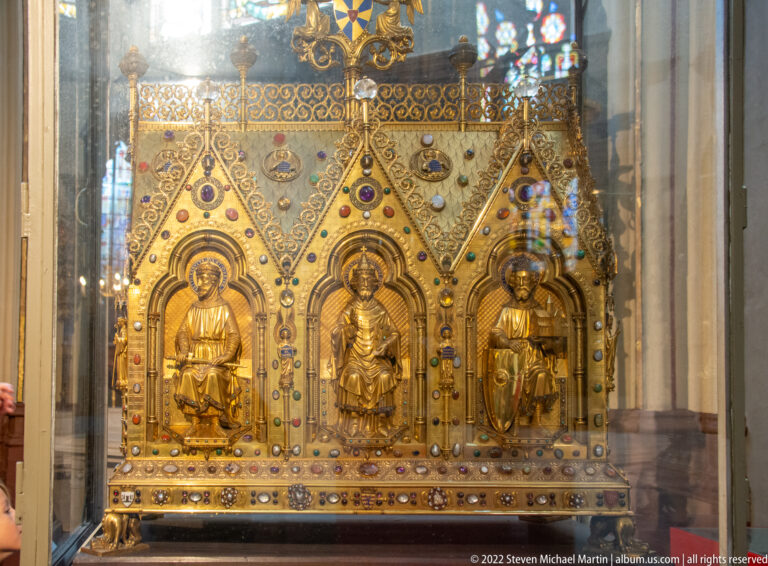 Gilded reliquary with three saint figures in pointed niches, jeweled decoration, and Gothic gables behind glass.