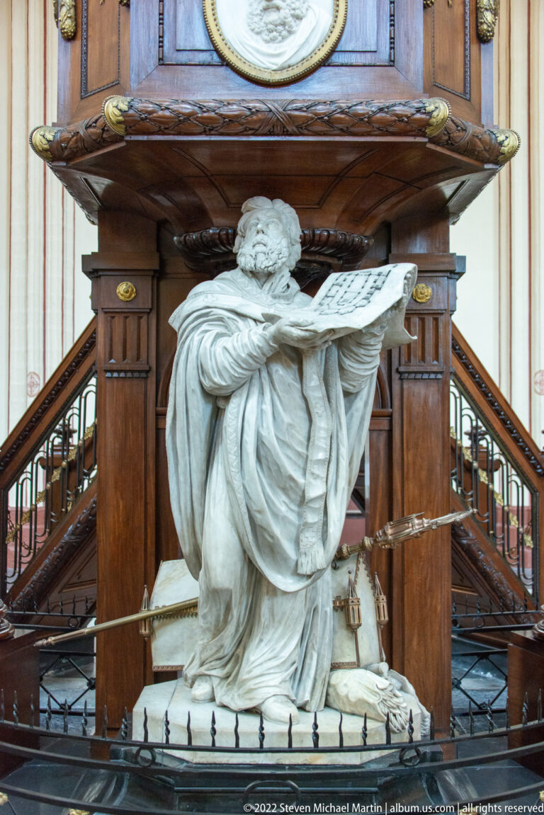 Kneeling stone figure holding an open book beneath the dark carved wooden base of a church pulpit.