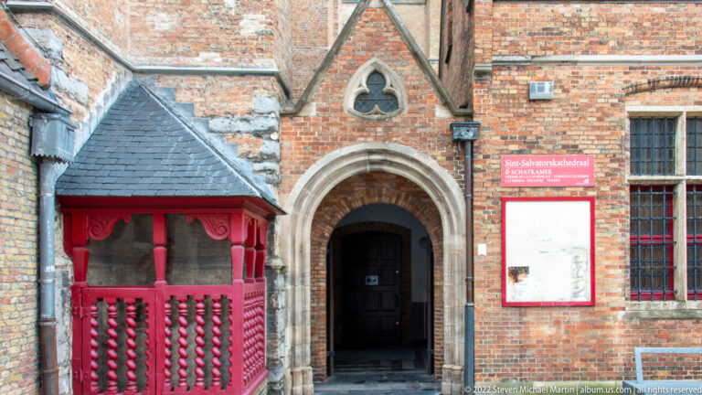 Pointed cathedral doorway beside a red timber porch, trefoil window, and Sint-Salvatorskathedraal visitor sign in Bruges.
