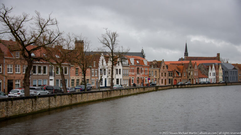Historic townhouses with varied gables and red roofs line a broad canal in Bruges beneath an overcast sky.