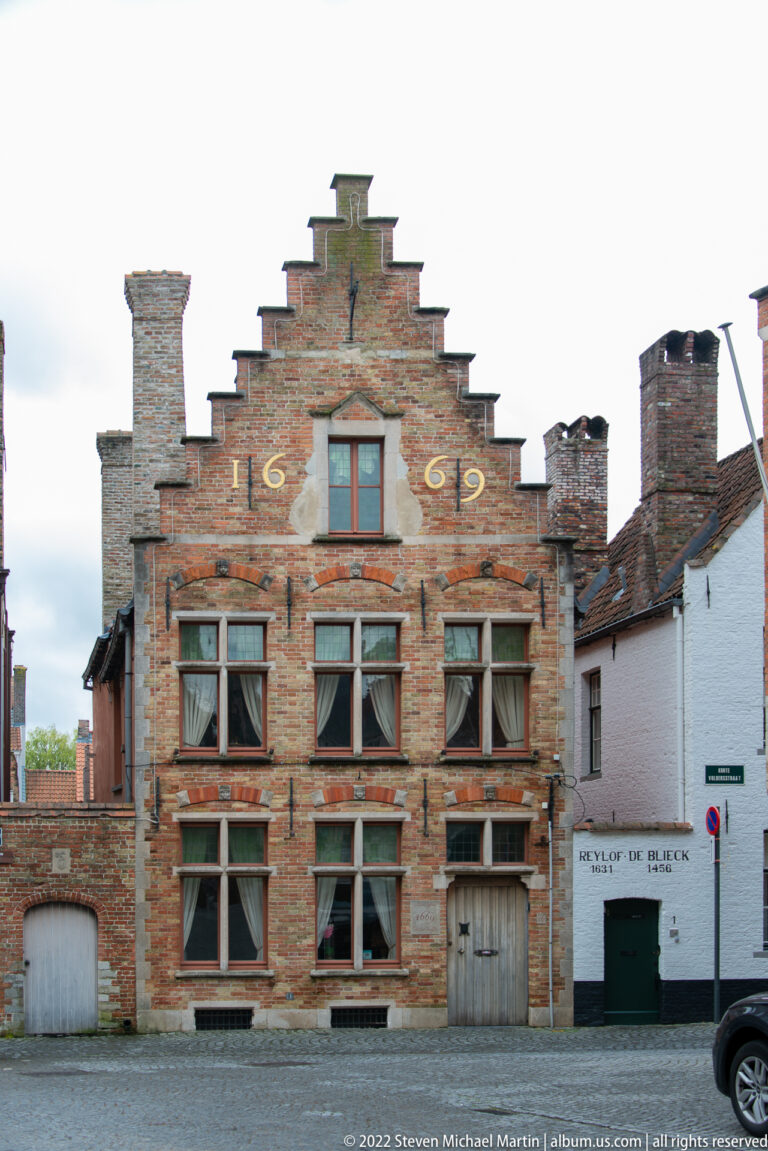 Weathered brick stepped-gable house dated 1609 with stone-framed windows, two doors, and tall chimneys in Bruges.