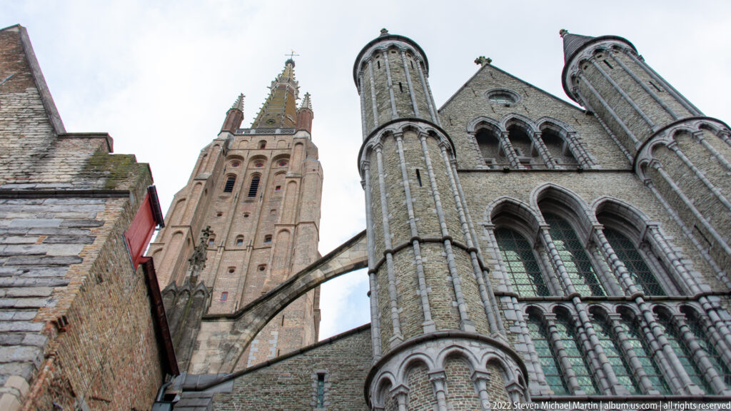 Stone church façade with paired round turrets and tall arched windows, with a brick tower rising behind in Bruges.