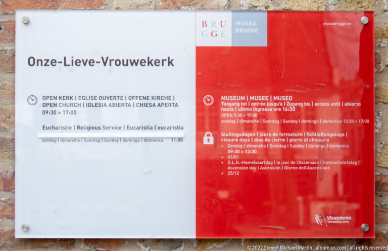 Red-and-white visitor board for Onze-Lieve-Vrouwekerk with church and museum opening hours in several languages.