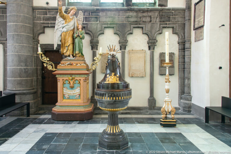 Polychrome saint statues and a tall ornate candle stand arranged against a dark stone church wall in Bruges.
