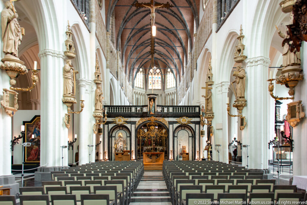 Long nave of the Church of Our Lady in Bruges with rib vaults, pointed arches, statues, and central rows of chairs.