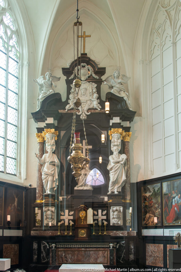 Dark side altar with pale sculpted figures, central crucifix, gilded details, and hanging lamps inside a Bruges church.