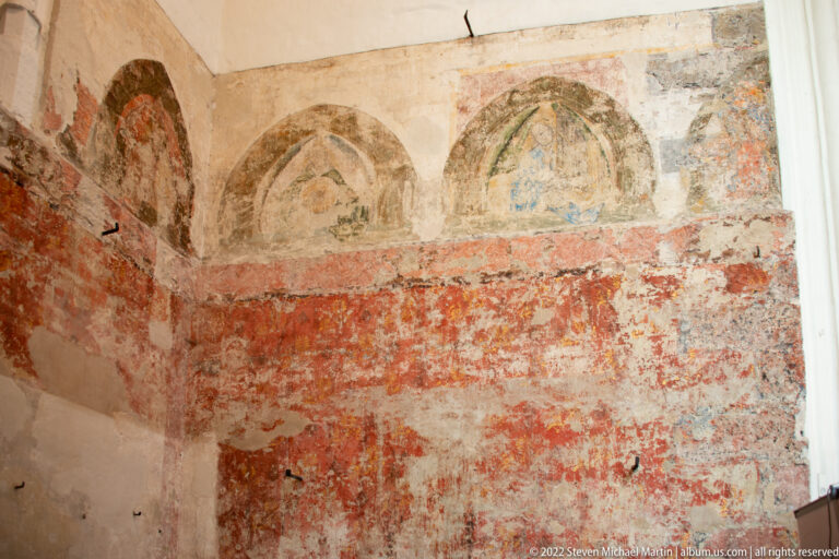 Faded medieval wall paintings with pointed-arch frames and red pigment surviving on worn plaster inside the church.