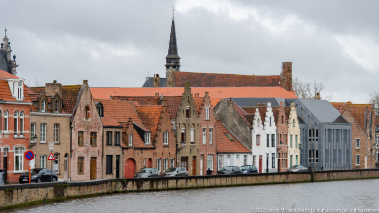 Gabled historic houses line a Bruges canal with a church roof and slender spire rising behind them.