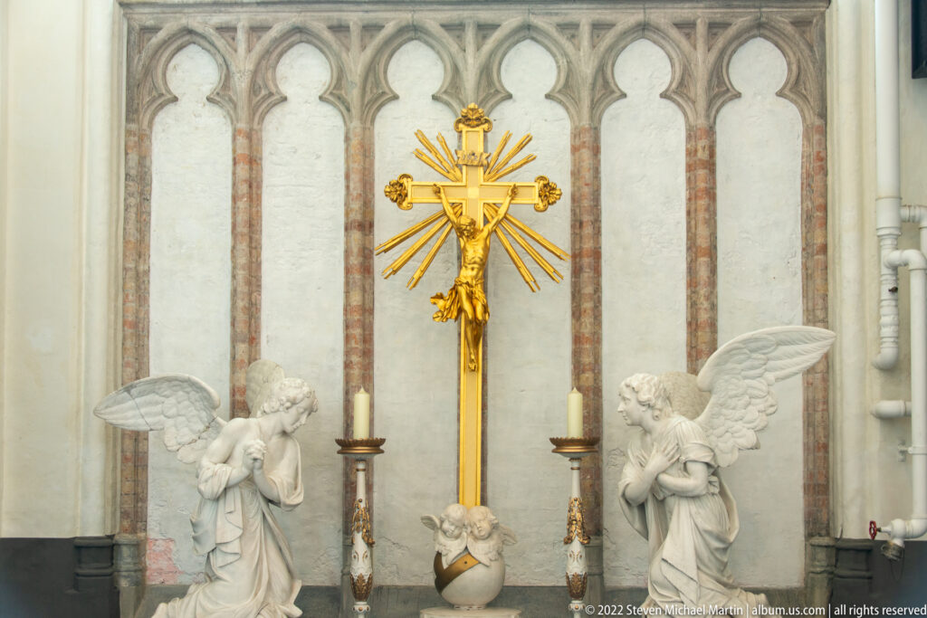 Gilded crucifix with radiant beams between two kneeling angel sculptures and candlesticks against a pale church wall.