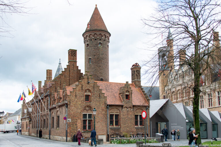 Historic brick complex with round conical-roofed tower, stepped gables, flags, and adjoining institutional buildings in Bruges.