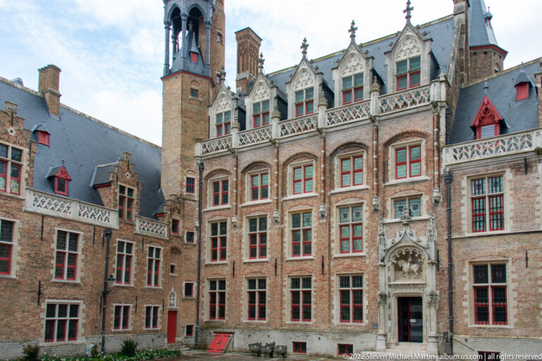 Ornate brick courtyard façade with pale stone tracery, pointed gables, red windows, and carved central doorway in Bruges.