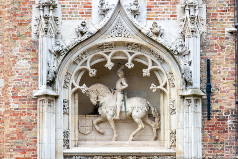 Stone relief of an armored rider on horseback beneath an ornate Gothic canopy set into a red-brick wall.