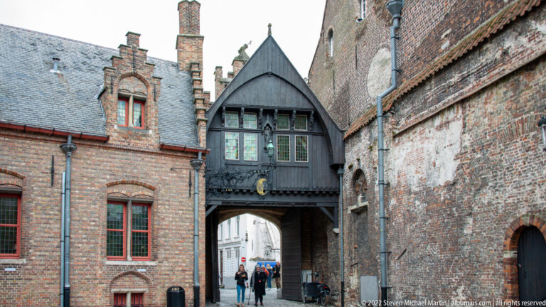 Dark timber upper passage bridges a narrow Bruges lane between old brick buildings with red-trimmed windows.