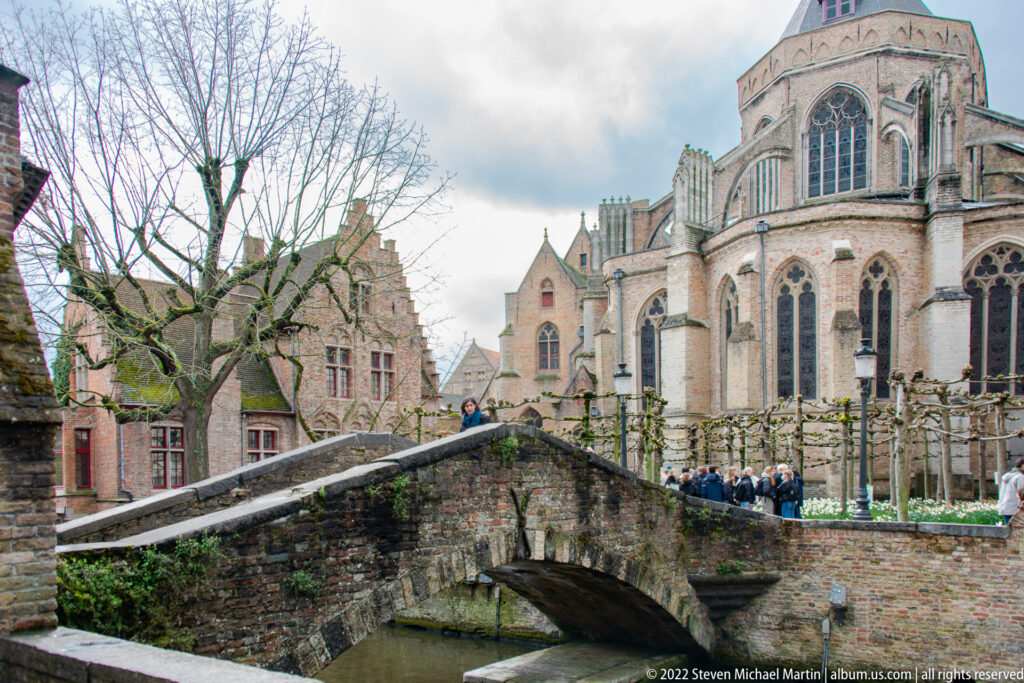 Arched brick footbridge over a Bruges canal with the large Church of Our Lady rising behind and visitors nearby.