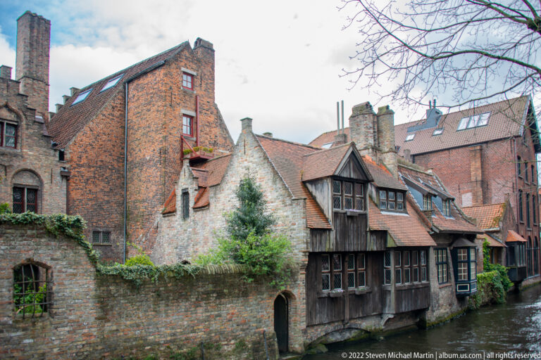 Weathered brick and timber houses with steep roofs and chimneys crowd the edge of a narrow Bruges canal.