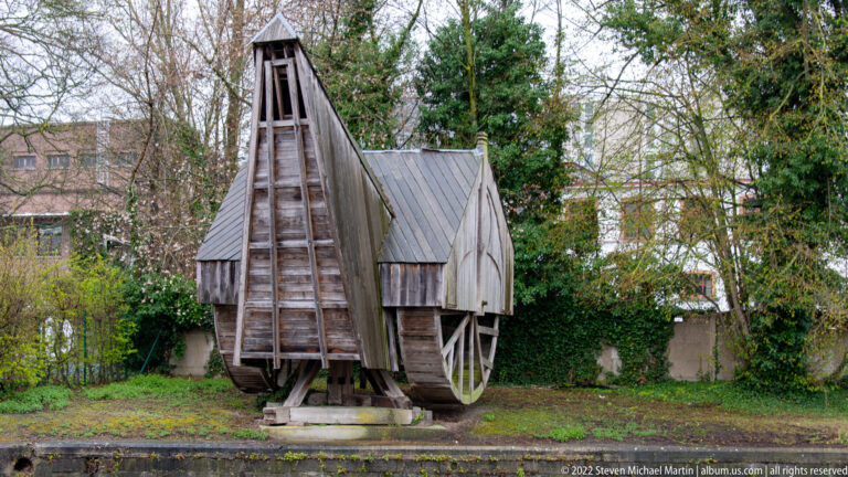 Weathered timber mechanism with large side wheels and tall wooden frame beside a Bruges waterway.