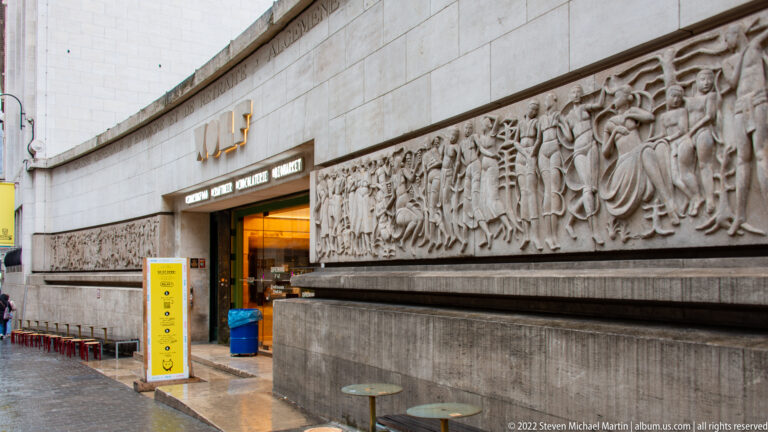 Long pale stone relief of stylized human figures beside a recessed entrance in a modern Brussels building.