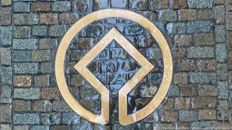Brass-colored World Heritage emblem embedded in dark cobblestone paving.