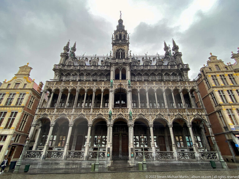 Low-angle view of the Maison du Roi with arcades, elaborate Gothic tracery, pinnacles, and central tower under dark clouds.