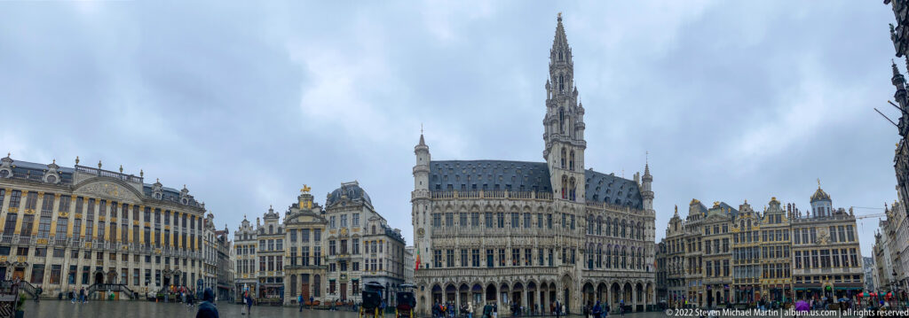 Wide view of Brussels Town Hall and neighboring guildhouses across the wet Grand-Place under an overcast sky.