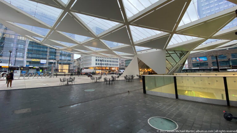 Modern Brussels railway-station concourse beneath an angular glass canopy, with travelers, bicycles, signs, and glazed building fronts.
