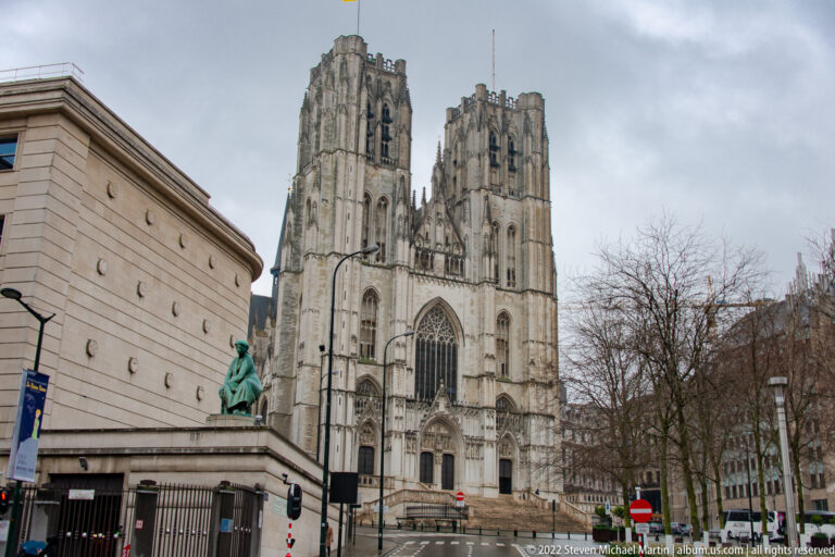 Street-level view of Brussels’ Cathedral of St. Michael and St. Gudula, with twin towers, Gothic portal, trees, and road signs.
