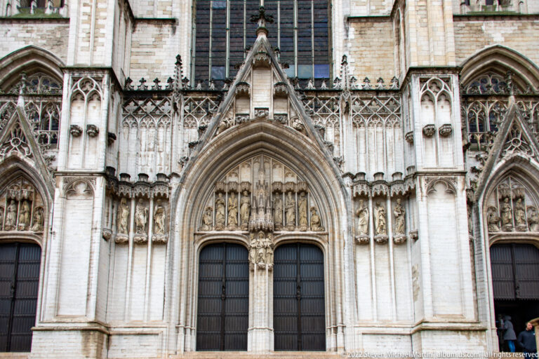 Frontal close view of a Gothic cathedral portal with pointed archivolts, carved figures, tracery, and dark double doors.