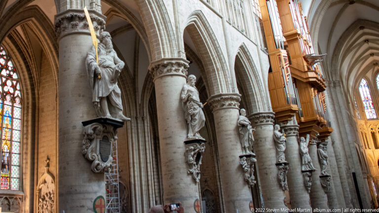 Stone apostle statues mounted on large cathedral columns beside pointed arches, with organ casework visible farther down the nave.