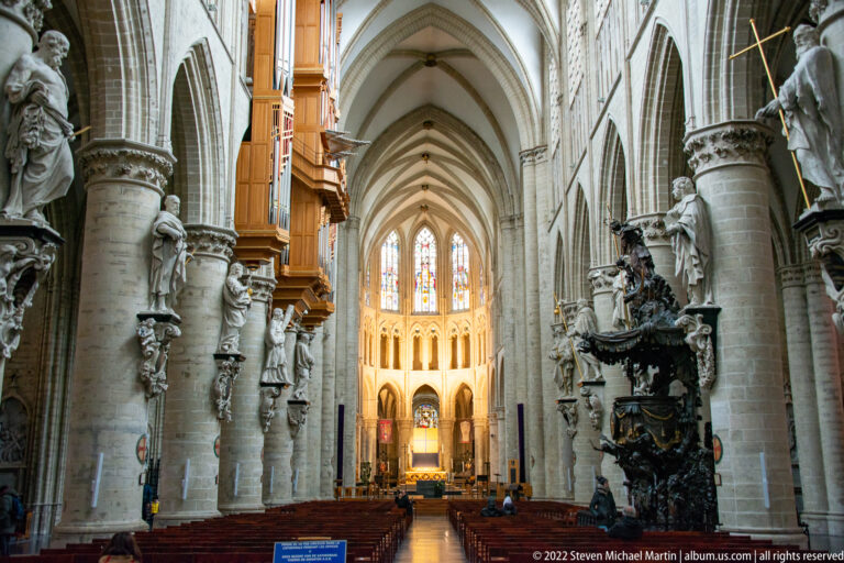 Centered view down the cathedral nave with pointed arches, ribbed vaults, column statues, pews, and the dark Baroque pulpit at right.