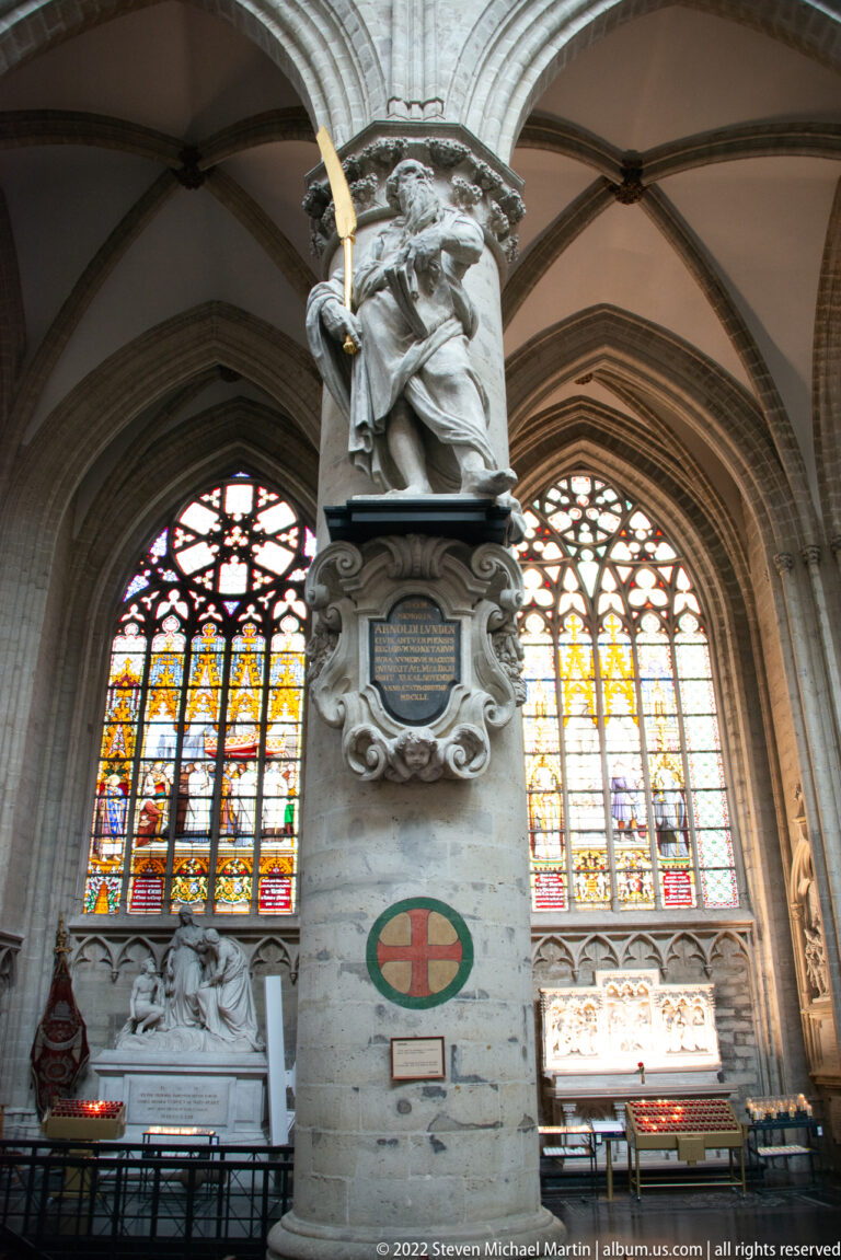 Apostle statue on a carved column bracket with a gilded attribute, set against tall multicolored stained-glass windows.
