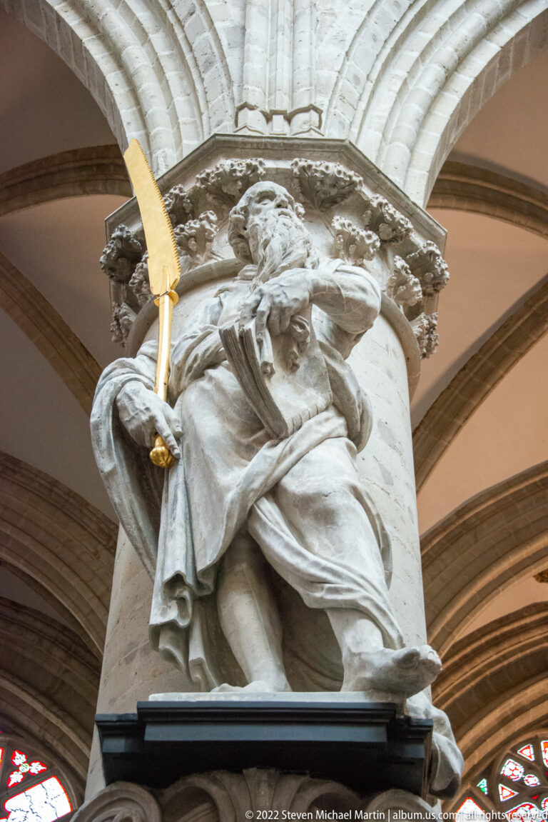 Close-up of a pale stone apostle statue on a column bracket holding a long gilded blade-like attribute.
