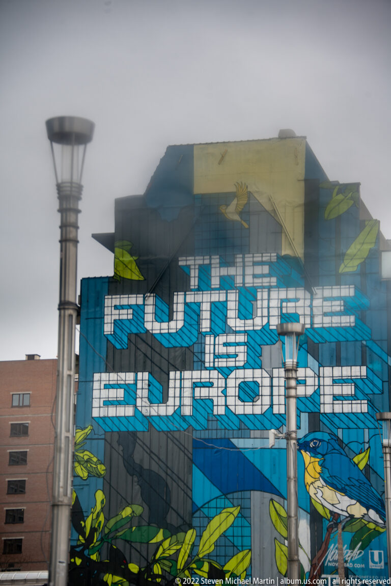 Large Brussels wall mural with blue-and-white FUTURE lettering, green foliage, and colorful bird imagery beside a streetlamp.