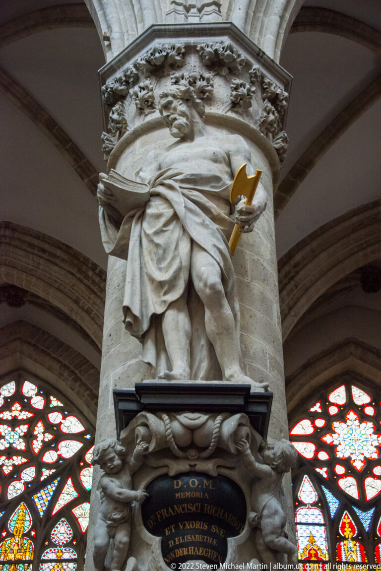 Standing stone figure on an ornate cathedral memorial above a dark oval inscription panel, with stained glass behind.