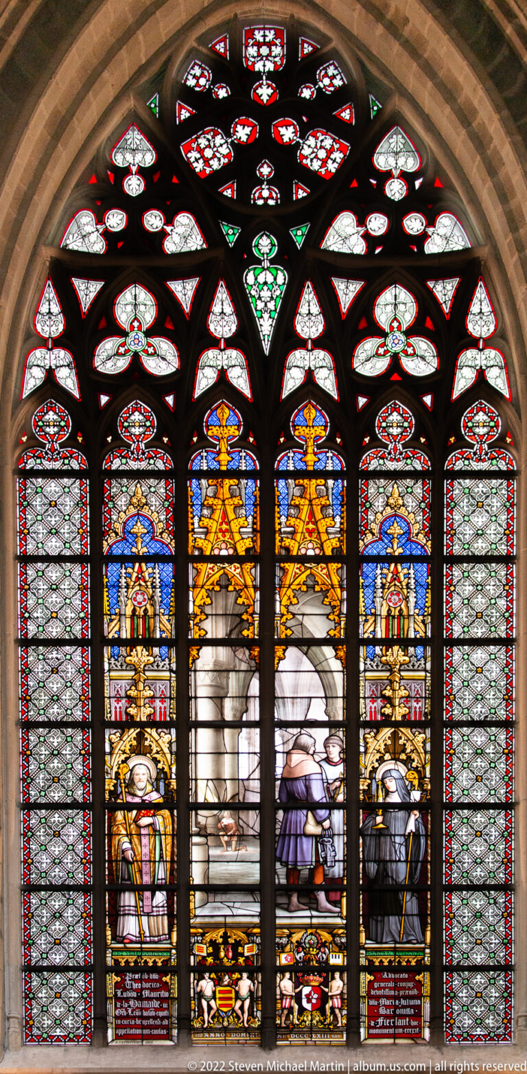 Arched stained-glass window with clerical figures, central narrative scenes, heraldic details, and small tracery lights above.