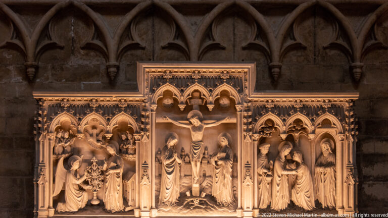 Illuminated carved altarpiece with the Crucifixion in the center and smaller Passion scenes beneath Gothic pointed arches.