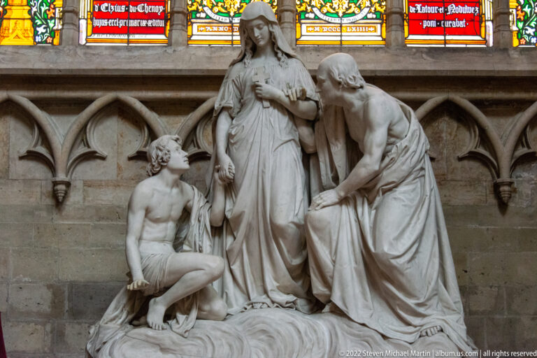 White devotional sculpture of the Virgin and Child with kneeling and seated figures beneath stained-glass windows.