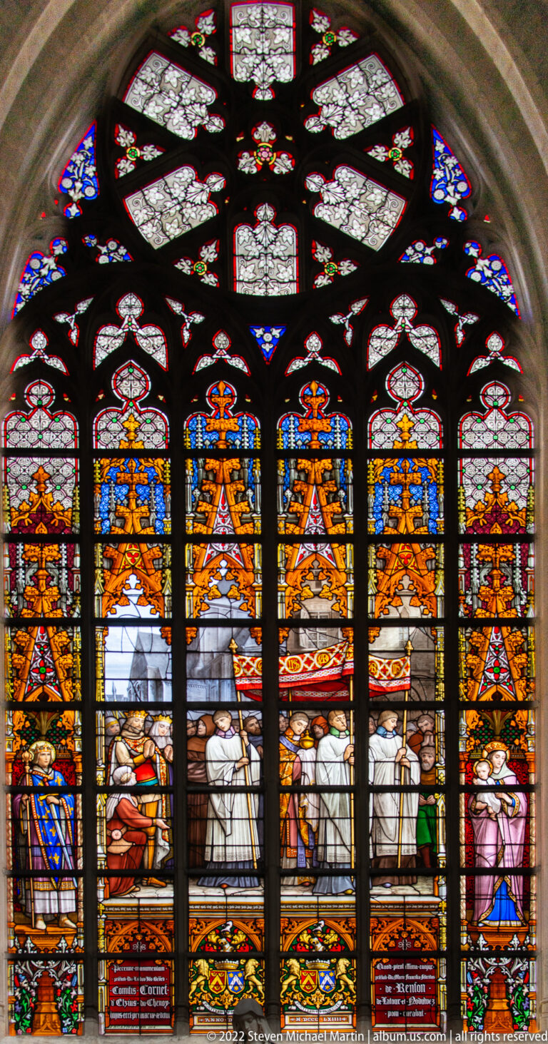 Five-lancet stained-glass window with richly dressed figures gathered beneath golden canopies and patterned tracery.