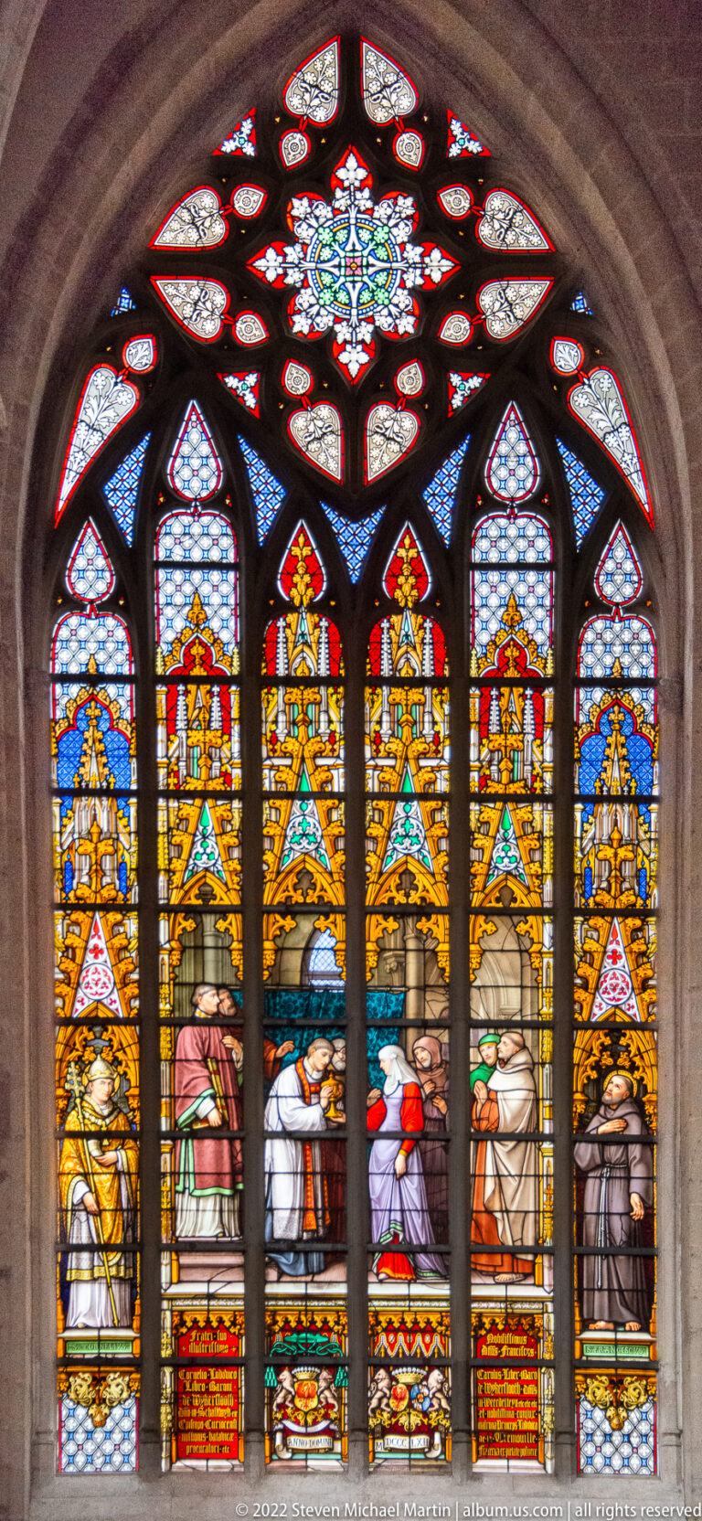 Five-lancet stained-glass window with kneeling and standing figures beneath gold canopies, crowned by a floral tracery light.