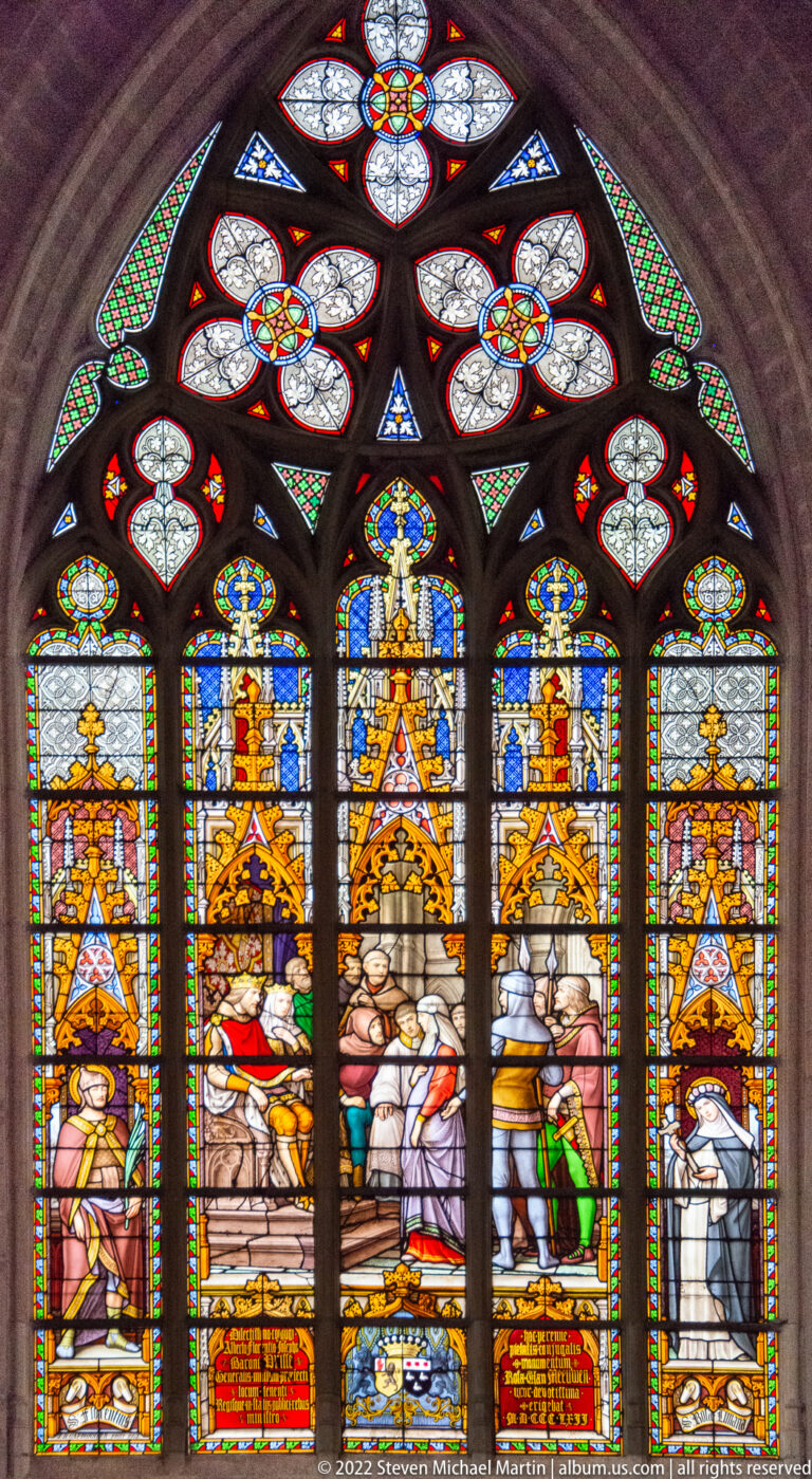 Arched stained-glass window with a crowded central scene, standing side figures, ornate canopies, and small upper tracery lights.