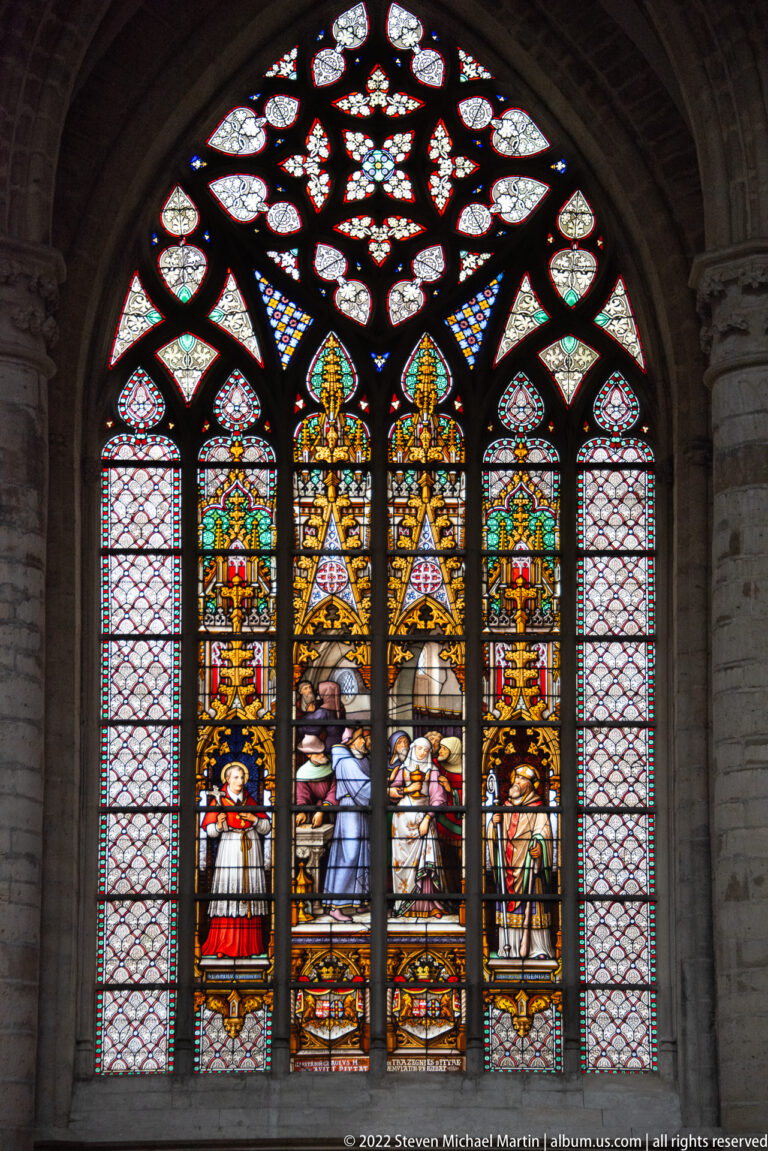 Arched stained-glass window with central figures beneath gold canopies, flanking figures, pale side panels, and rosette tracery.