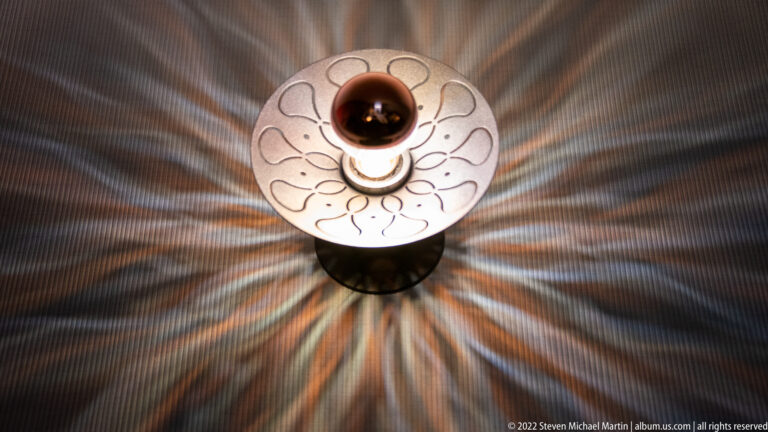 Circular decorative wall sconce with looping linework casts radiating bands of light across a dark ribbed wall.