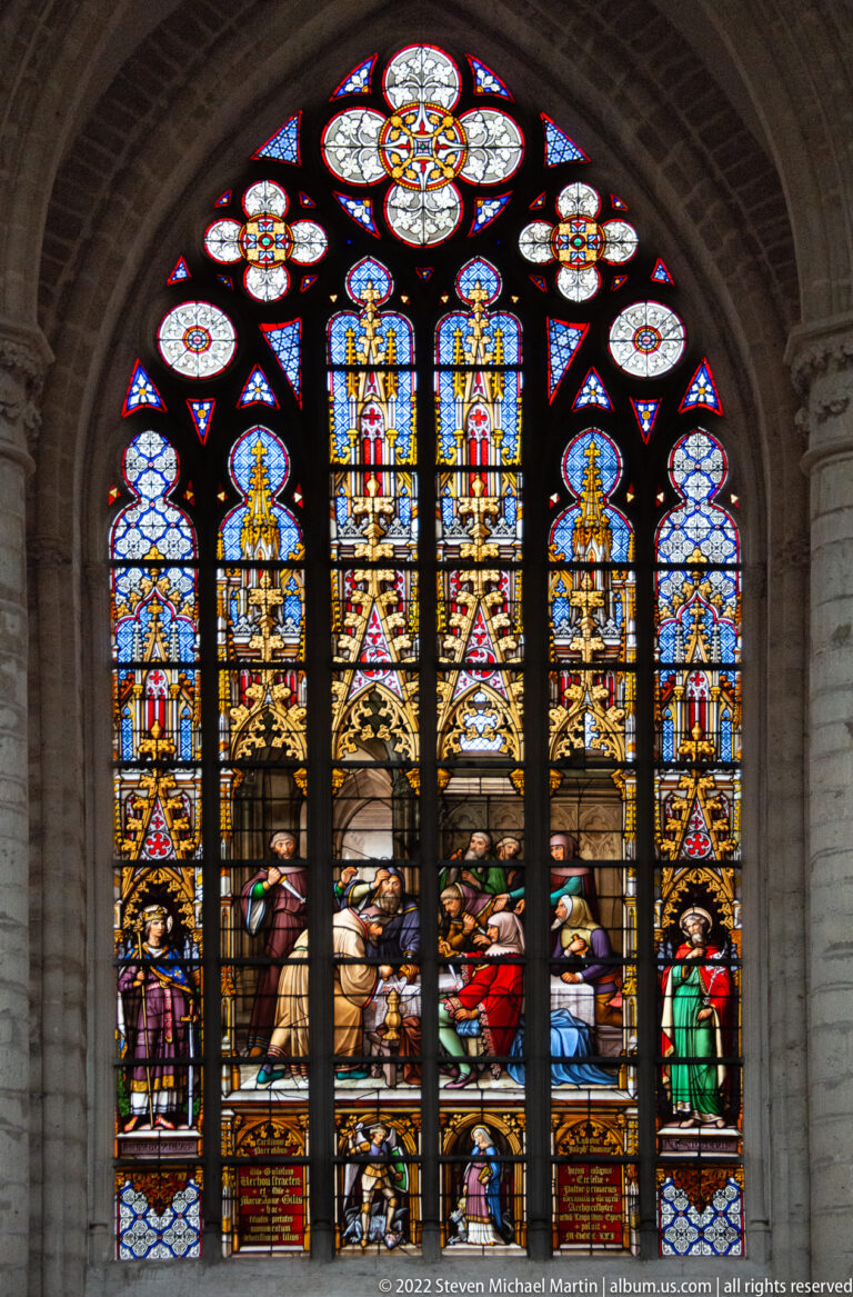 Arched stained-glass window with kneeling and standing figures below, two tall central figures, and a floral tracery light above.