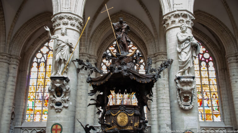 Dark monumental Baroque pulpit with elaborate figures and canopy between pale Gothic columns and stained-glass windows.