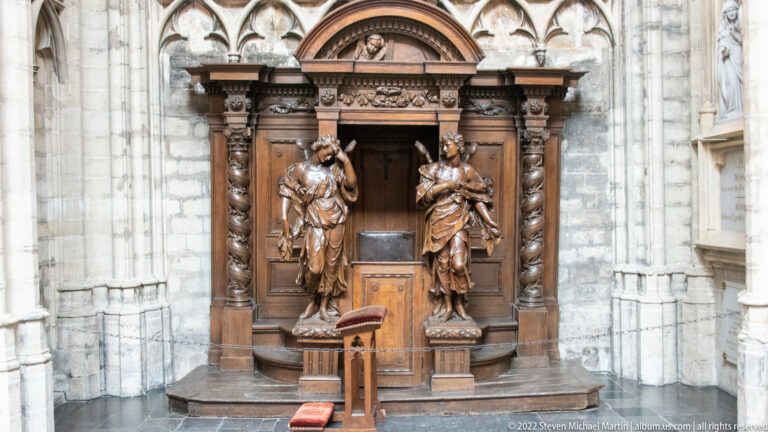 Frontal view of an ornate wooden confessional with two standing carved figures, columns, and a small kneeler against pale stone.