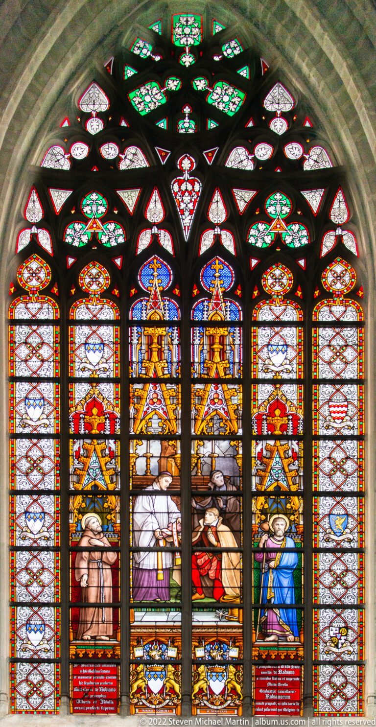 Five-lancet stained-glass window with kneeling figures in the center, gold canopies, pale side panels, and green upper tracery.