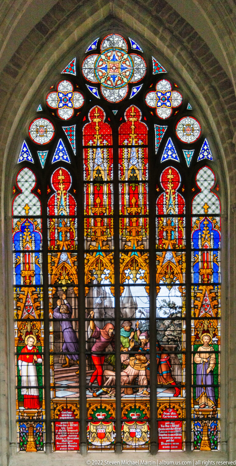 Arched stained-glass window with vivid red upper panels, crowded central figures, side saints, and floral tracery.