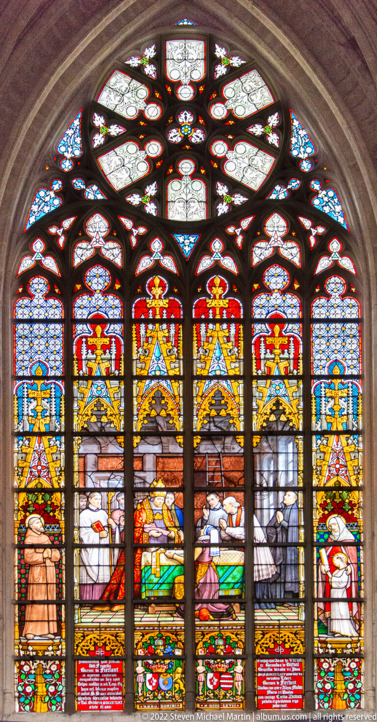 Arched stained-glass window with a recumbent figure surrounded by attendants beneath gold canopies and red-blue upper panels.