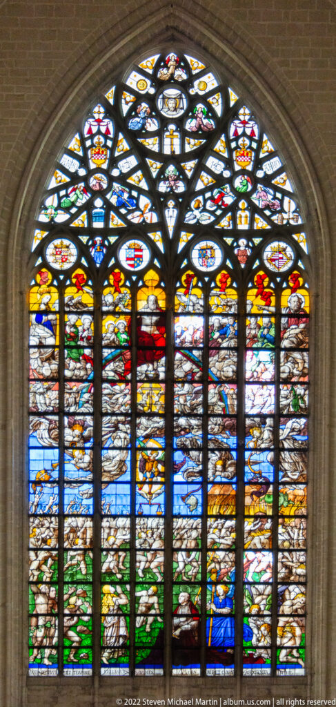 Five-lancet stained-glass landscape with small figures, buildings, green terrain, heraldic roundels, and a blue-robed figure at lower right.